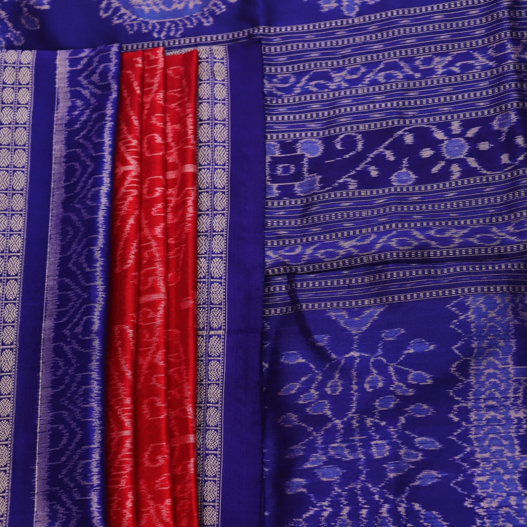 Close-up of red Sambalpuri silk saree fabric showing intricate woven motif texture and subtle blue threads.