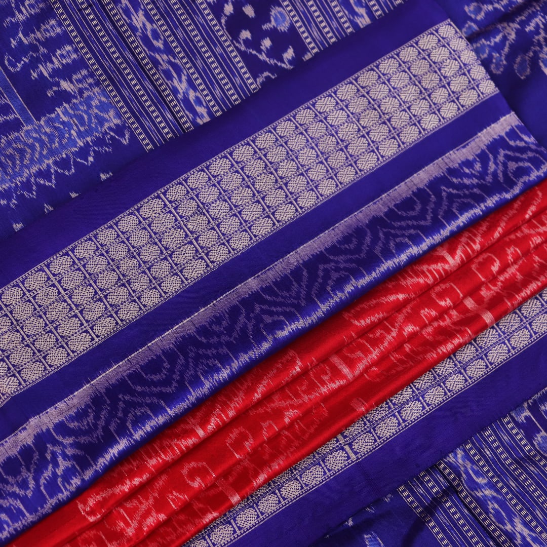 Close-up of red Sambalpuri silk saree fabric showing intricate woven motif texture and subtle blue threads.